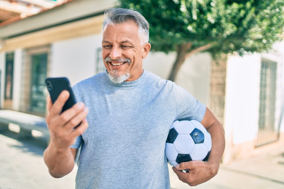 Mature Hispanic man holding a soccer ball while using phone